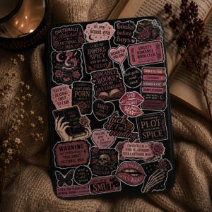 Op de afbeelding: Een zwarte e-reader hoes bedekt met roze en zwarte stickers met zinnen als "Smut" en "Romance Books". De stickers bevatten rozen, lippen en boekgerelateerde afbeeldingen. De hoes ligt op een getextureerd, beige oppervlak met een boek en een kaars.