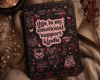May include: A black e-reader case featuring a pink floral and celestial pattern with the text 'this is my emotional support kindle', 'The Smutty Book Club', 'Morally Grey', and 'you had me at TRIGGER WARNING' alongside illustrations of a skull and books.