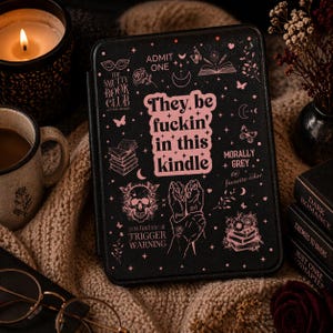 May include: Black Kindle cover with pink graphic text that reads "They be fuckin' in this kindle." The cover is decorated with pink illustrations of skulls, books, butterflies, and other designs. The cover is on a neutral-colored knitted surface.