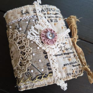 May include: A fabric-covered journal featuring a beige linen exterior with printed French text including LA BAJOU and PARIS. The cover is embellished with a circular crochet doily, a white lace strip, a dusty pink fabric flower with a button, and yellow stitching.