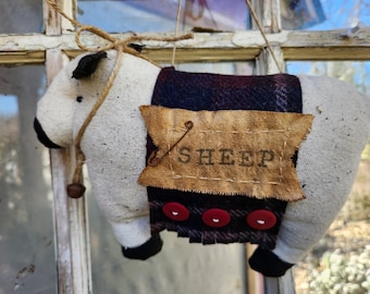 May include: A white fabric sheep ornament with black eyes and hooves. The sheep is wearing a plaid fabric scarf with red buttons and a brown fabric tag that says "SHEEP".