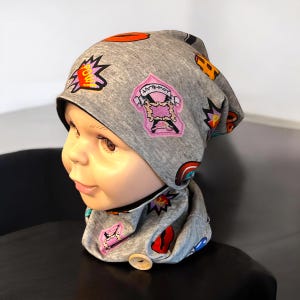 Può includere: Set di berretto e scaldacollo grigi con toppe colorate in stile fumetto. Le toppe includono la parola "POW!" in un'esplosione gialla, una toppa rosa con un teschio e un arcobaleno. Lo scaldacollo ha una chiusura con bottone in legno.