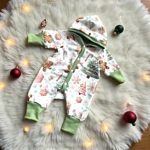 Op de afbeelding: Een witte baby-onesie met kerstthema, met peperkoekmannetjes, kerstbomen en sneeuwvlokken. De onesie heeft groene randen op de capuchon, manchetten en langs de knoopsluiting. De onesie is dichtgeknoopt en ligt op een witte, pluizige ondergrond.