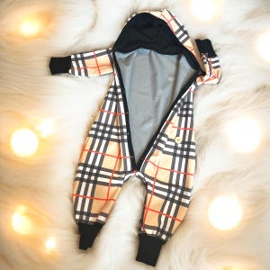 Può includere: Tuta da bambino a quadri beige, rossi, neri e bianchi con cappuccio e polsini neri. La tuta ha una chiusura con bottoni sul davanti ed è realizzata in materiale morbido. L'indumento è progettato per neonati e bambini piccoli.