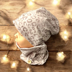 Pode incluir: Um gorro bege e um cachecol combinando com padrão de leopardo. O gorro tem uma aba dobrável e o cachecol tem um fecho de botão.