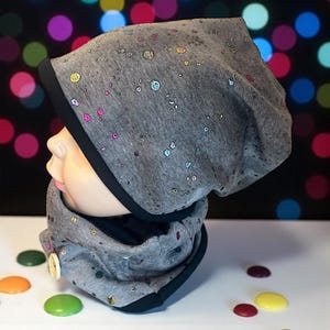 Puede incluir: Conjunto de gorro y braga de cuello grises con ribete negro y un patrón de puntos iridiscentes de colores. La braga de cuello tiene un cierre de botón. El conjunto se muestra sobre una cabeza de maniquí sobre un fondo borroso de luces de colores.