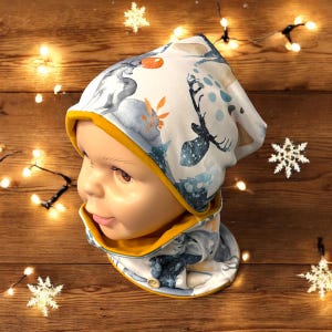 Pode incluir: Um conjunto de gorro e cachecol de inverno temático de inverno branco e azul com acabamento amarelo. O gorro apresenta um veado, um coelho e outras criaturas da floresta. O cachecol tem um fecho de botão.