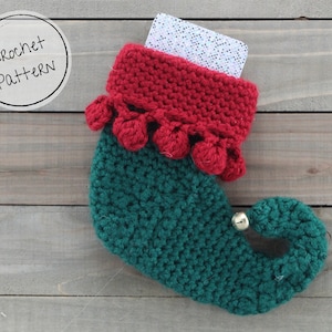Puede incluir: Un calcetín de elfo de crochet verde y rojo con una campana en la punta y pompones alrededor del puño. El calcetín contiene una tarjeta de regalo.