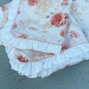 May include: A folded white blanket featuring a floral design of peach and red roses. The blanket has a ruffled edge with a pink trim. The fabric appears soft and delicate, suitable for bedding or decorative use.