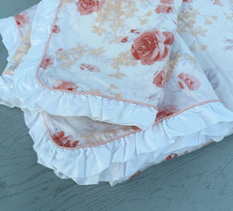 May include: A folded white blanket featuring a floral design of peach and red roses. The blanket has a ruffled edge with a pink trim. The fabric appears soft and delicate, suitable for bedding or decorative use.