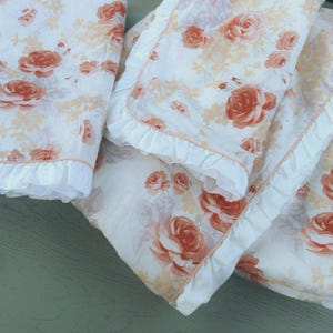 May include: Bedding set featuring a floral rose design in peach and cream hues. The set includes a blanket and pillowcase with frilled edges. The roses are large and detailed, creating a romantic and classic aesthetic.