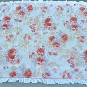 May include: A white blanket featuring a floral design of coral and peach roses and small peach flowers. It has a ruffled white border. The blanket is suitable for a bed or cot.