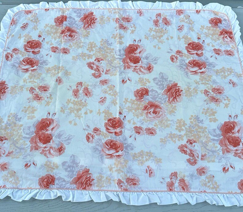 May include: A white blanket featuring a floral design of coral and peach roses and small peach flowers. It has a ruffled white border. The blanket is suitable for a bed or cot.