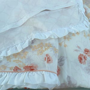 May include: Close-up of a floral patterned bedding set. The set features a white background with large red and peach roses, and smaller yellow flowers. The edges of the bedding are trimmed with white frills.