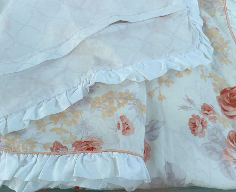 May include: Close-up of a floral patterned bedding set. The set features a white background with large red and peach roses, and smaller yellow flowers. The edges of the bedding are trimmed with white frills.