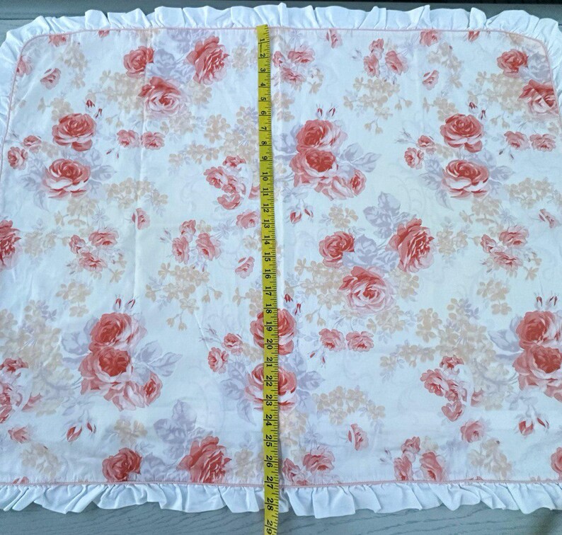 May include: A rectangular fabric featuring a floral pattern with a white base and pink roses and peach-colored flowers. The fabric is approximately 74 cm long, with a ruffled edge.