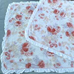 May include: Two white baby blankets with a floral rose pattern in shades of peach and pink. Each blanket has a white ruffled edge. The blankets are folded and layered on top of each other.