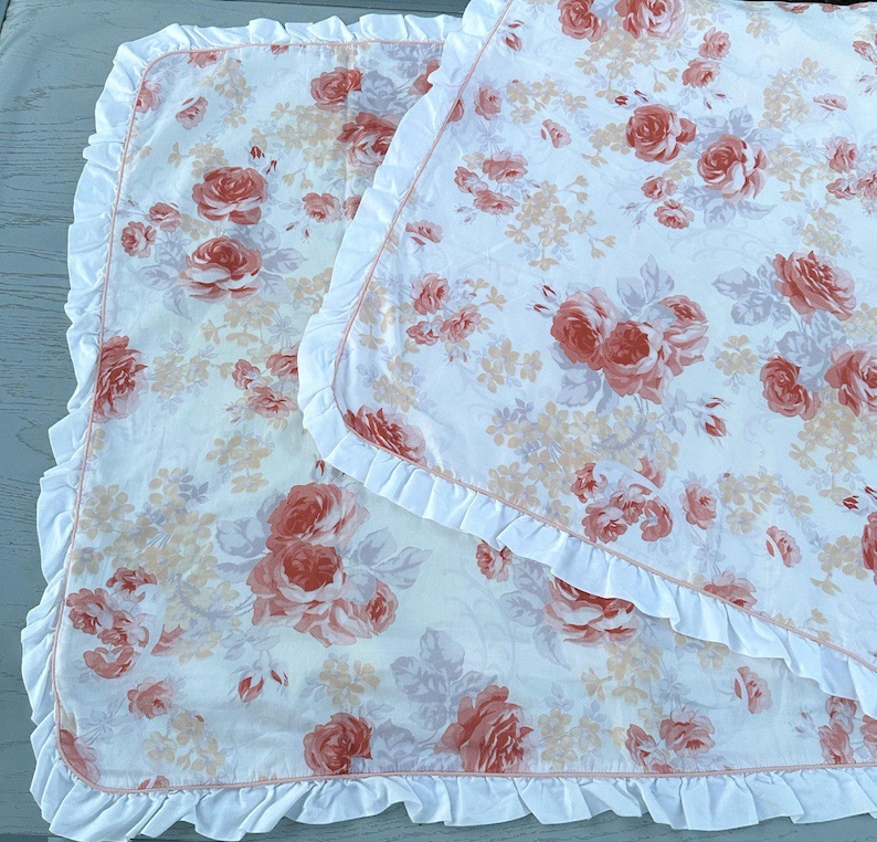May include: Two white baby blankets with a floral rose pattern in shades of peach and pink. Each blanket has a white ruffled edge. The blankets are folded and layered on top of each other.