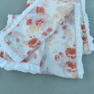 May include: A floral bedding set with a white base, adorned with coral roses and peach-toned flowers. The set has a ruffled edge and a pink trim. The bedding is designed with a romantic floral pattern.