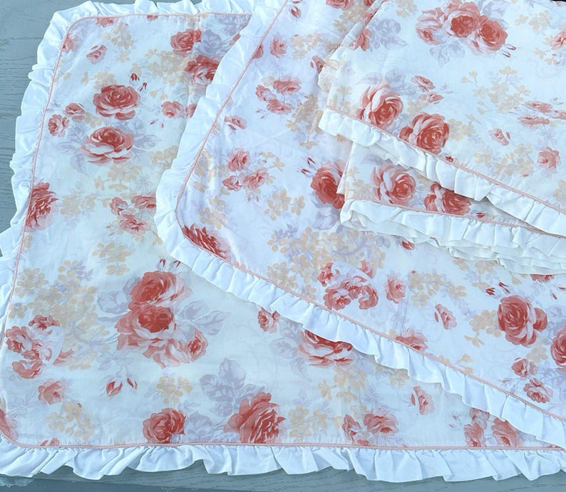 May include: White bedding set featuring a rose floral pattern in peach and pink hues. The set includes a blanket and pillowcases, all finished with a ruffled edge.