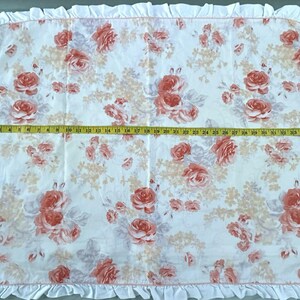 May include: A white floral blanket with a ruffled edge. The blanket features a pattern of red and pink roses and other flowers. The blanket measures approximately 89 cm wide.