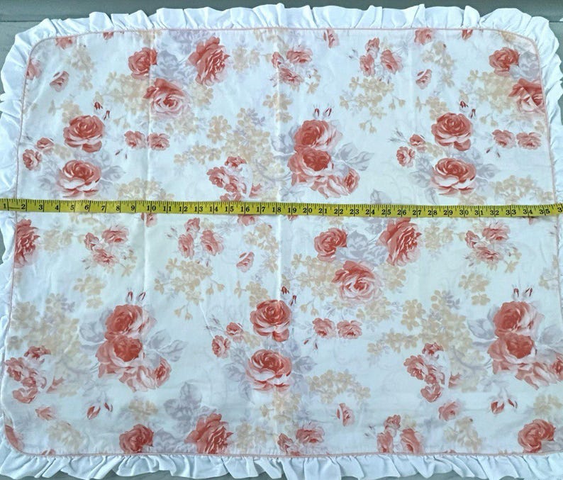 May include: A white floral blanket with a ruffled edge. The blanket features a pattern of red and pink roses and other flowers. The blanket measures approximately 89 cm wide.