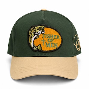 May include: A dark green baseball cap with a tan brim and a top button. The front features an oval patch with a yellow background, a green fish illustration, a small cross, and the embroidered text "FISHER OF MEN". A gold logo is on the side.