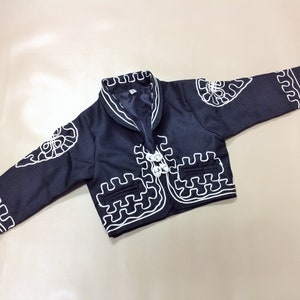 Puede incluir: Una chaqueta negra con detalles bordados en blanco. La chaqueta tiene un dise&ntilde;o tradicional mexicano con cuello de pico y cierre de bot&oacute;n.