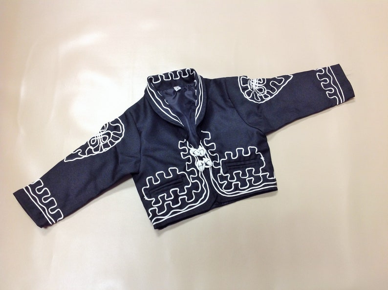 Puede incluir: Una chaqueta negra con detalles bordados en blanco. La chaqueta tiene un dise&ntilde;o tradicional mexicano con cuello de pico y cierre de bot&oacute;n.
