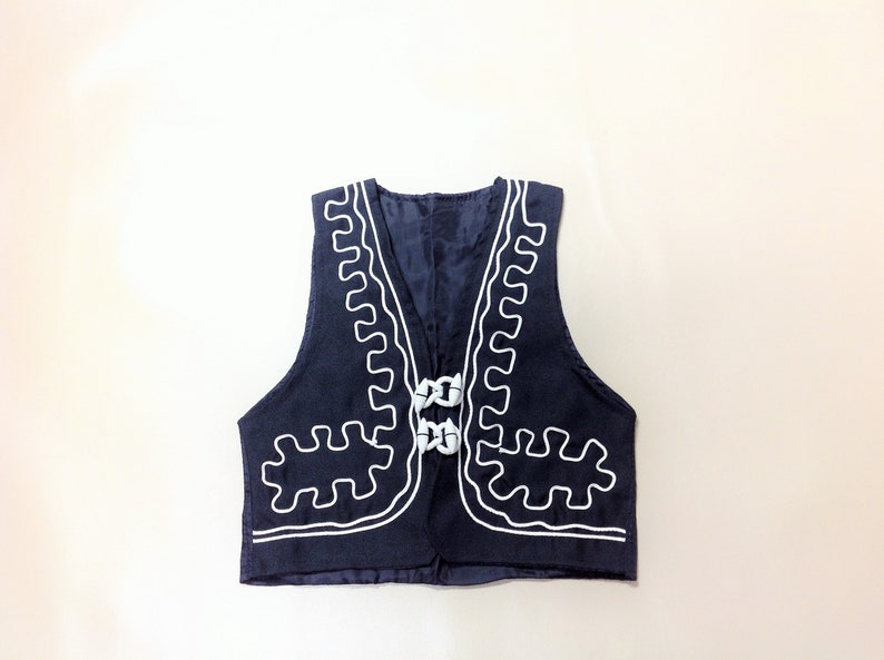Puede incluir: Un chaleco azul marino con detalles bordados en blanco alrededor del cuello y la parte inferior. El chaleco tiene cuello en V y dos botones blancos.