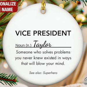 May include: A white ceramic Christmas ornament with gold trim, featuring the text "VICE PRESIDENT" and a definition. The name "Taylor" is written below. The ornament is personalized and includes the phrase "See also: Superhero."
