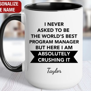 May include: White ceramic mug with a black rim and handle. The mug has black text that reads "I never asked to be the world's best program manager but here I am absolutely crushing it" with the name "Taylor" below the text.