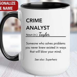 May include: White ceramic mug with a black handle and rim. The mug features the text "CRIME ANALYST" and a definition, with the name "Taylor" and the phrase "See also: Superhero". The top left corner has the text "PERSONALIZE THE NAME".