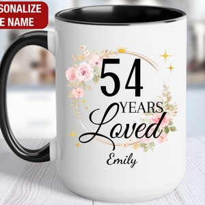 May include: White ceramic mug with a black rim and handle. The mug features a floral design with a gold circle around the number 54 and the text "YEARS Loved". The name "Emily" is printed below the text.