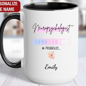 May include: White coffee mug with a black handle and black rim. The mug has a pink and blue progress bar with the text "Neuropsychologist in progress..." and the name "Emily" at the bottom.