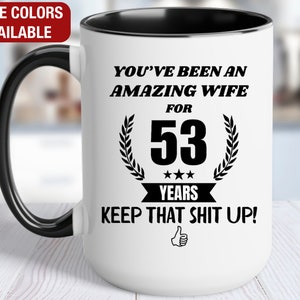 May include: White ceramic mug with a black interior and handle. The mug features the text "YOU'VE BEEN AN AMAZING WIFE FOR 53 YEARS KEEP THAT SHIT UP!" in black, with a thumbs-up graphic. The words "MORE COLORS AVAILABLE" are in a red box.