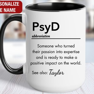 May include: White ceramic mug with a black handle and black rim. The mug has a black and white design that reads "PsyD abbreviation Someone who turned their passion into expertise and is ready to make a positive impact on the world. See also: Taylor"