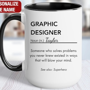 May include: White ceramic mug with a black handle and black rim. The mug has a graphic design that reads "GRAPHIC DESIGNER Noun (n.) Taylor Someone who solves problems you never knew existed in ways that will blow your mind. See also: Superhero"