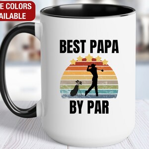 May include: White ceramic coffee mug with a black rim and handle. The mug features a retro-style sunset graphic with a golfer and the text "Best Papa By Par".