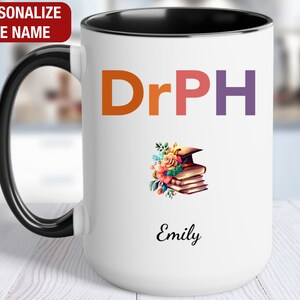 May include: White ceramic coffee mug with a black handle and black rim. The mug has the text "DrPH" in colorful letters and a floral design with a graduation cap and books. The name "Emily" is written below the design.