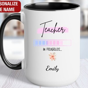 May include: White ceramic mug with a black handle and black rim. The mug has a pink and blue progress bar with the text "Teacher in progress..." and the name "Emily" at the bottom.