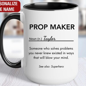 May include: White ceramic mug with a black rim and handle. The mug has black text that reads "PROP MAKER" and a definition of the word "PROP MAKER" with the name "Taylor" inserted. The text reads "Noun (n.) Taylor Someone who solves problems you never knew existed in ways that will blow your mind. See also: Superhero."