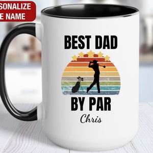 May include: White ceramic coffee mug with a black rim and handle. The mug features a retro-style graphic of a golfer with a sunset background. The text "Best Dad By Par" is printed in black, with the name "Chris" printed below.