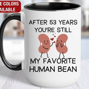 May include: White ceramic mug with a black handle and rim. The mug has a graphic of two red beans with faces and the text "After 53 years you're still my favorite human bean."