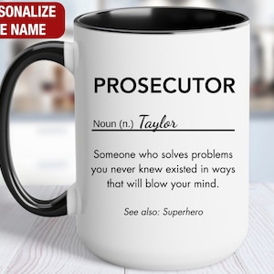 May include: White ceramic mug with a black handle and black rim. The mug has a definition of the word "Prosecutor" with the name "Taylor" inserted. The definition reads: "Someone who solves problems you never knew existed in ways that will blow your mind. See also: Superhero."