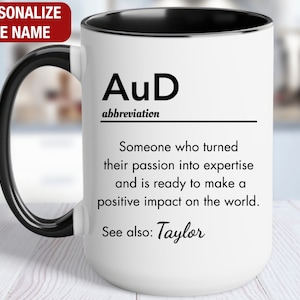 May include: White ceramic mug with a black handle and black rim. The mug has a black and white design that reads "AuD abbreviation Someone who turned their passion into expertise and is ready to make a positive impact on the world. See also: Taylor"
