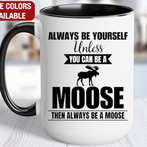 May include: White ceramic coffee mug with a black rim and handle. The mug features a black silhouette of a moose and the text "Always be yourself unless you can be a moose then always be a moose".
