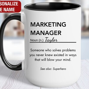 May include: White ceramic coffee mug with a black handle and black rim. The mug has a black and white design that reads "MARKETING MANAGER Noun (n.) Taylor Someone who solves problems you never knew existed in ways that will blow your mind. See also: Superhero"