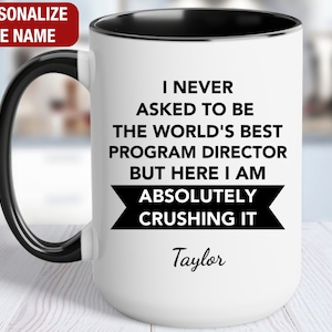 May include: White ceramic mug with a black rim and handle. The mug has black text that reads "I never asked to be the world's best program director but here I am absolutely crushing it." The name "Taylor" is written in cursive below the text.