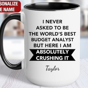 May include: White ceramic mug with a black handle and rim. The mug features the text "I NEVER ASKED TO BE THE WORLD'S BEST BUDGET ANALYST BUT HERE I AM ABSOLUTELY CRUSHING IT" and the name "Taylor".